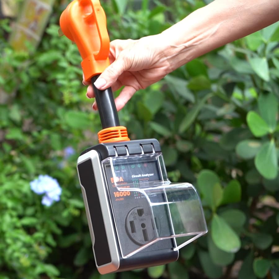 Hand holding a 50 amp RV surge protector with built-in circuit analysis againsts green foliage background